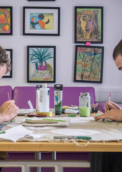 Picture of two people painting in an art class. Lots of works of art on the walls in the background